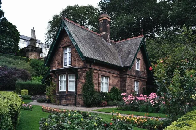 Design reference: brick cottage with a steep gabled slate roof and a tall chimney, set behind a rose garden and lawn