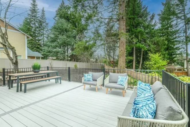 Design reference: a painted deck with a wood railing and outdoor lounge seating under tall evergreen trees, showing the railings and finish that need upkeep in a wet climate